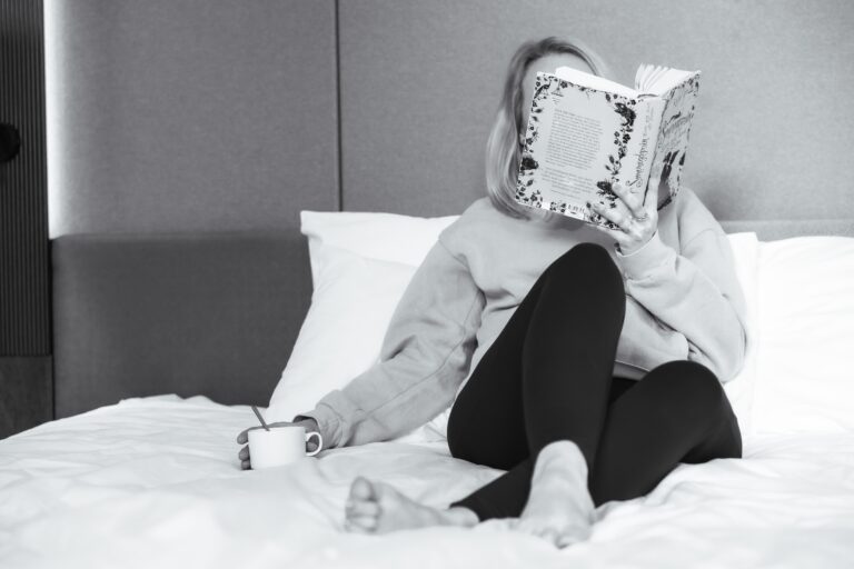 Femme assise sur un lit, le visage caché derrière un livre, dans un moment calme.